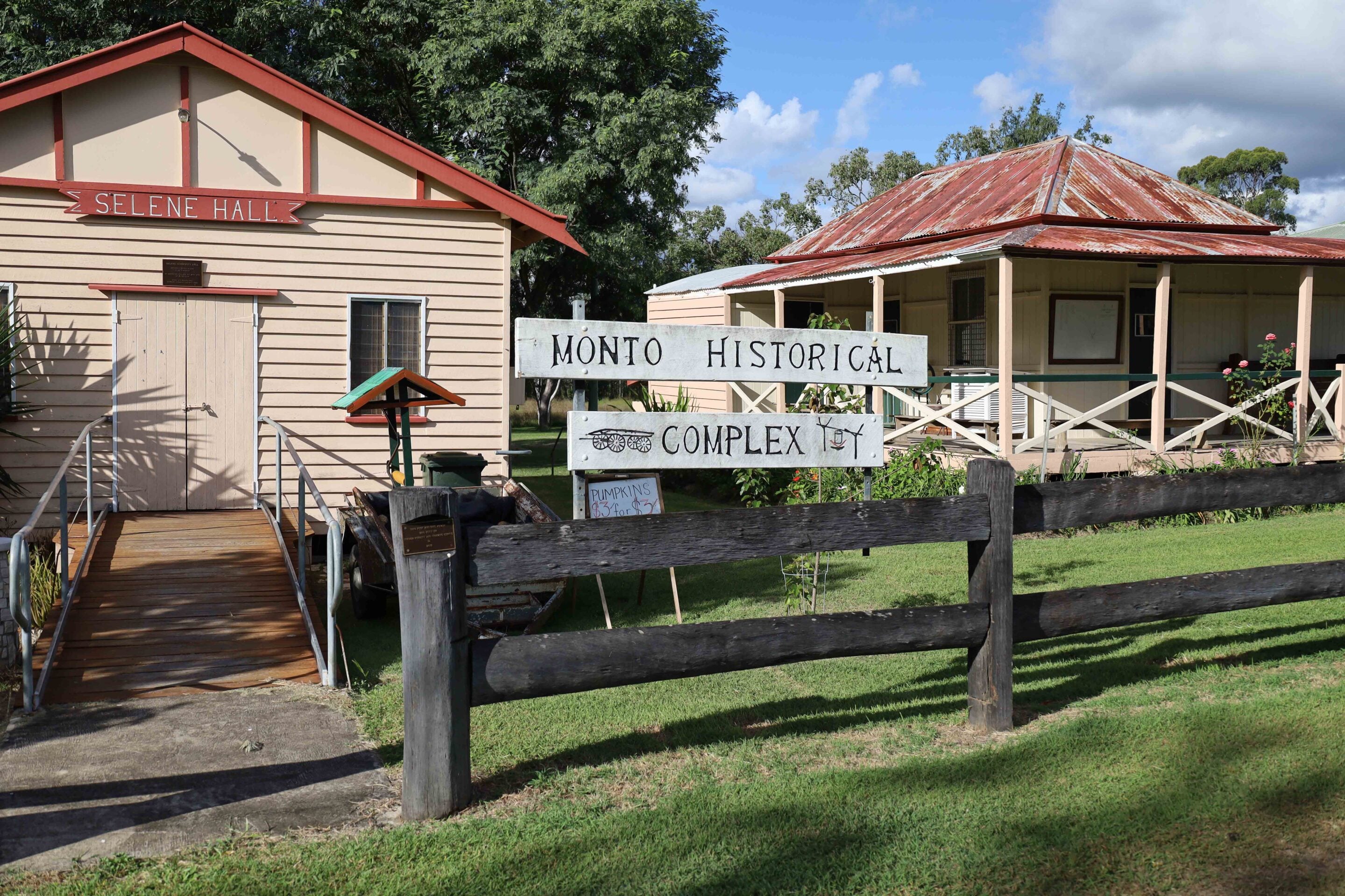 Historic wooden buildings with red roofs and a sign reading 'Monto Historical Complex' in a sunny yard.