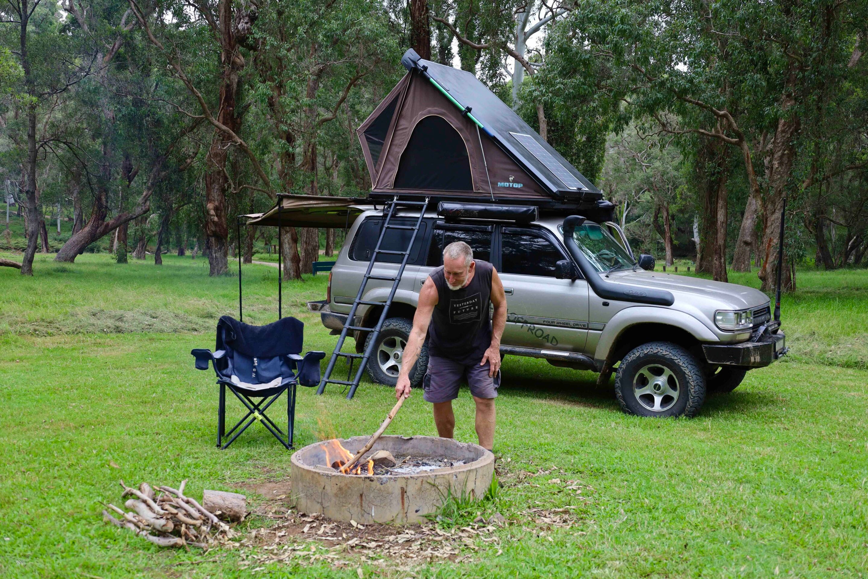 SUV with a rooftop tent parked in a forested campsite; man tends a campfire nearby.116 characters?