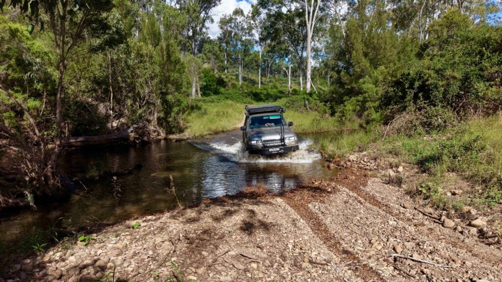 Silver SUV crossing a shallow river in a dense green forest, splashing water as it drives through.