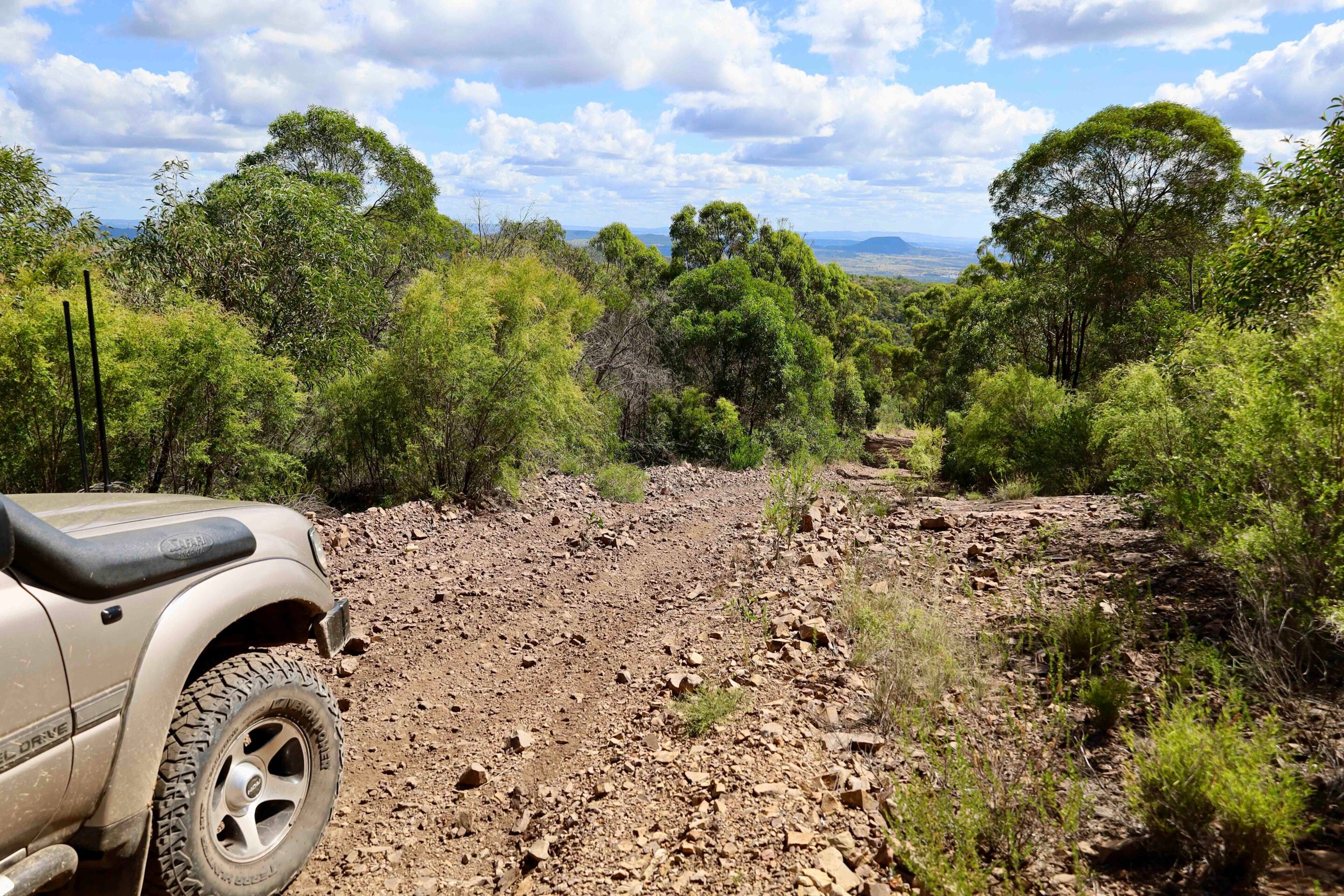 Beige off-road vehicle on a rocky dirt trail with green shrubs and a distant blue-sky landscape.