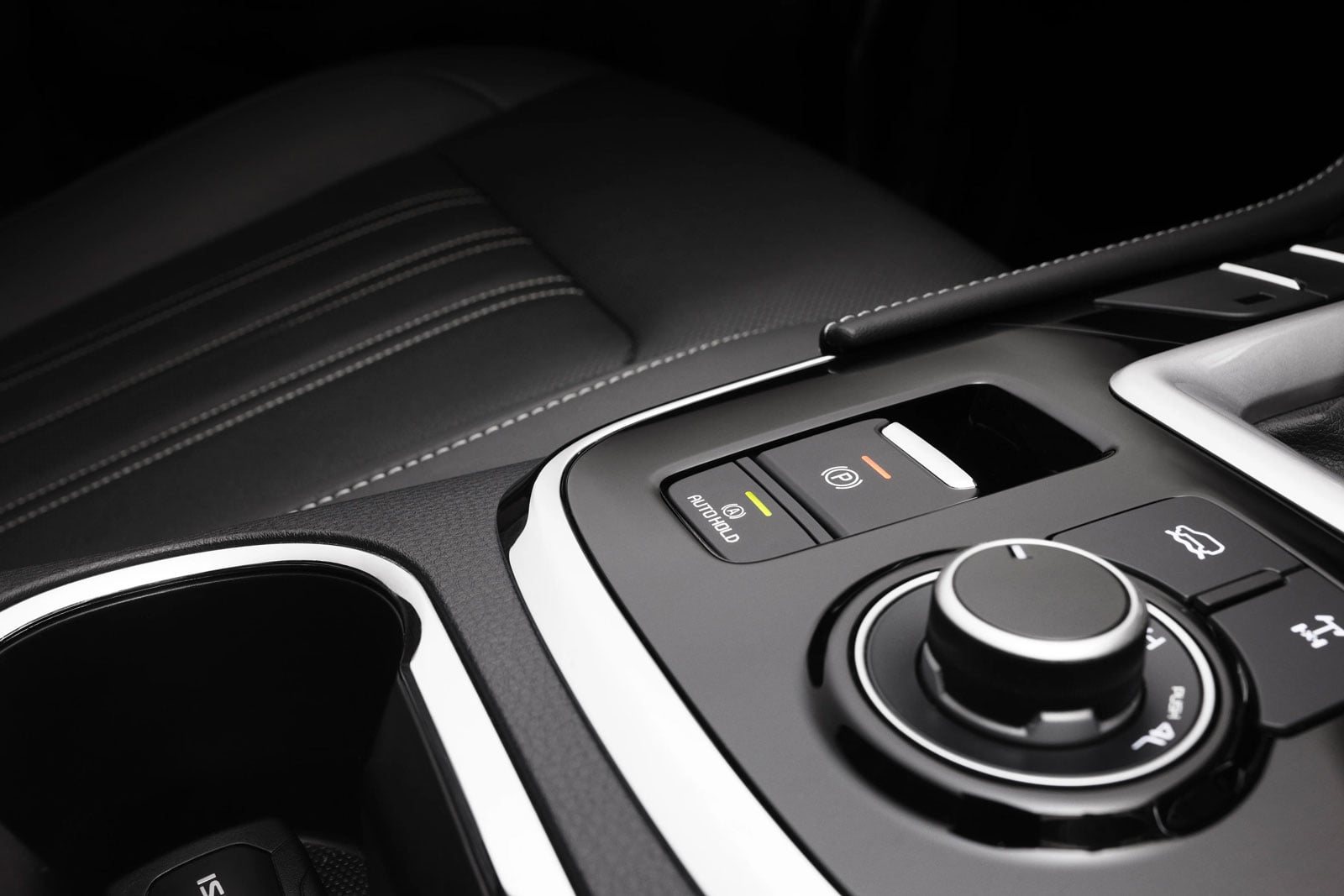 Close-up of a car's center console featuring the electronic parking brake button, a control dial, and surrounding glossy trim on black leather with white stitching.