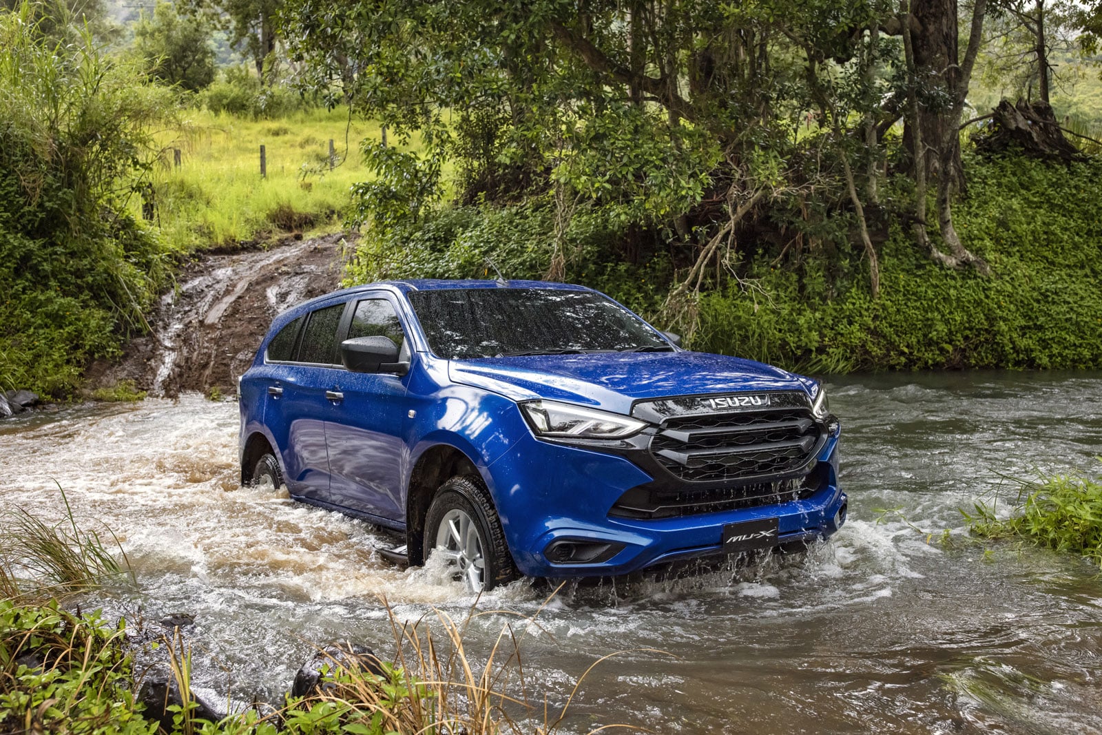 Blue Isuzu MU-X SUV crosses a shallow muddy river with green, trees and grass in the background.
