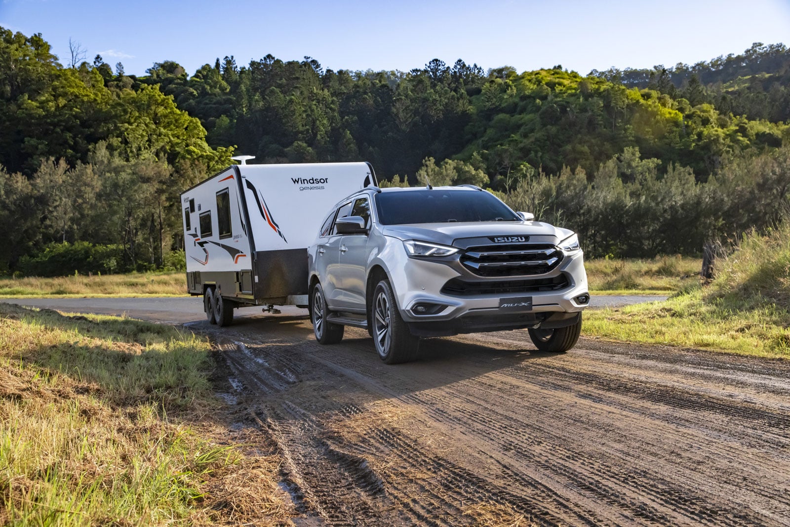 Isuzu MU-X SUV towing a white Windsor Genesis caravan along a dirt rural road with trees in the background.