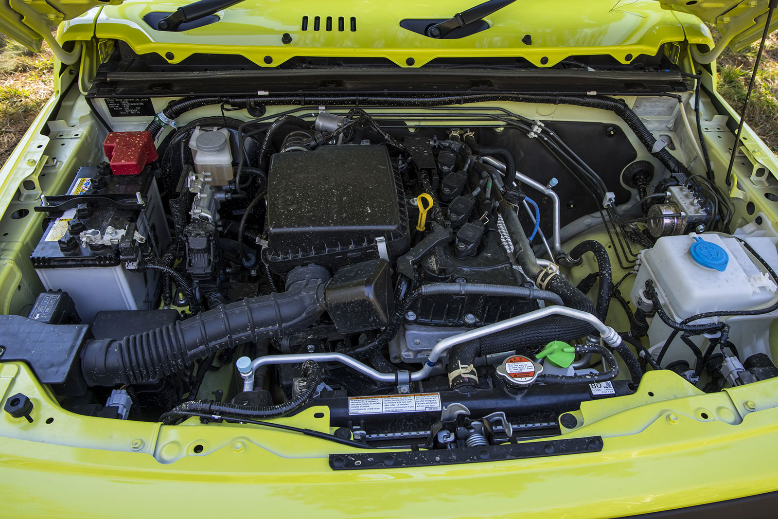 Open-hood view of a neon yellow car showing its engine bay, with the battery on the left, coolant reservoir on the right, and various hoses and wiring visible across the engine.