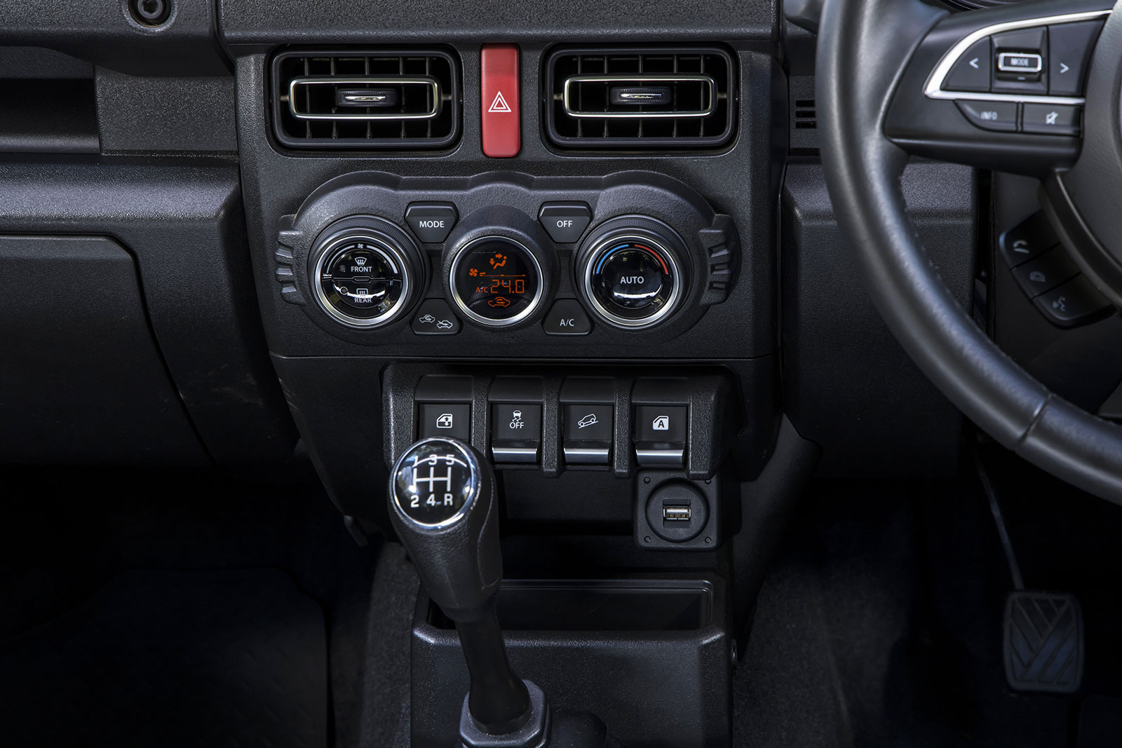 Center console of a car showing climate control dials, hazard button, and a manual gear shift (1-5 R) with surrounding switches and a USB port nearby.