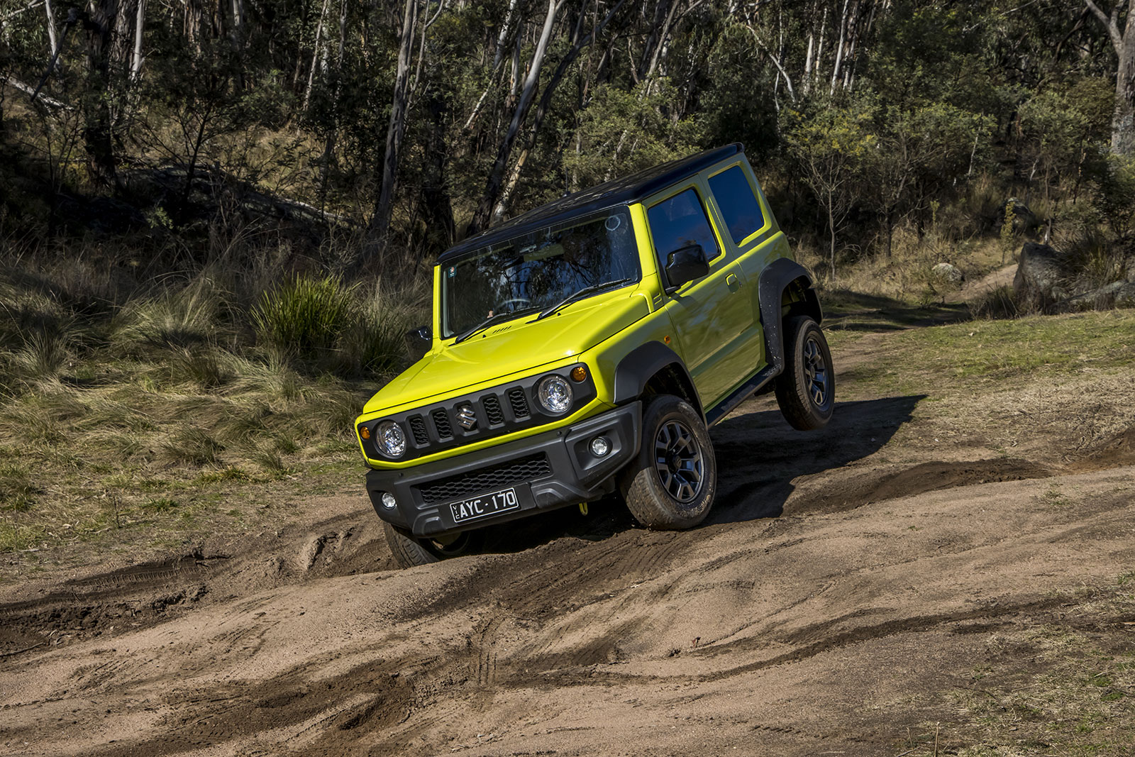 Bright yellow-green Suzuki Jimny climbs a rutted dirt trail in a forest, wheel lifted on uneven terrain