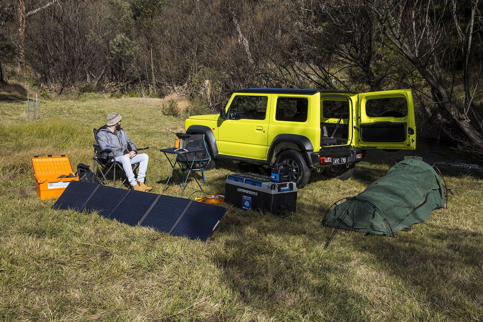 Man camping by a bright yellow off-road SUV with doors open, solar panel, and camping gear on a grassy field.