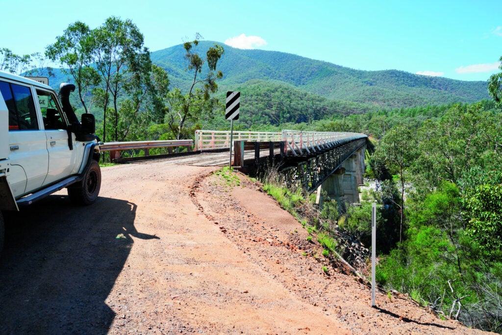 White off-road SUV parked on a dusty roadside near a metal bridge over a deep valley, with green hills in the distance and blue sky.