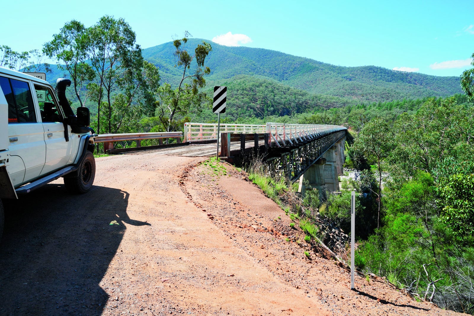 White off-road SUV parked on a dusty roadside near a metal bridge over a deep valley, with green hills in the distance and blue sky.