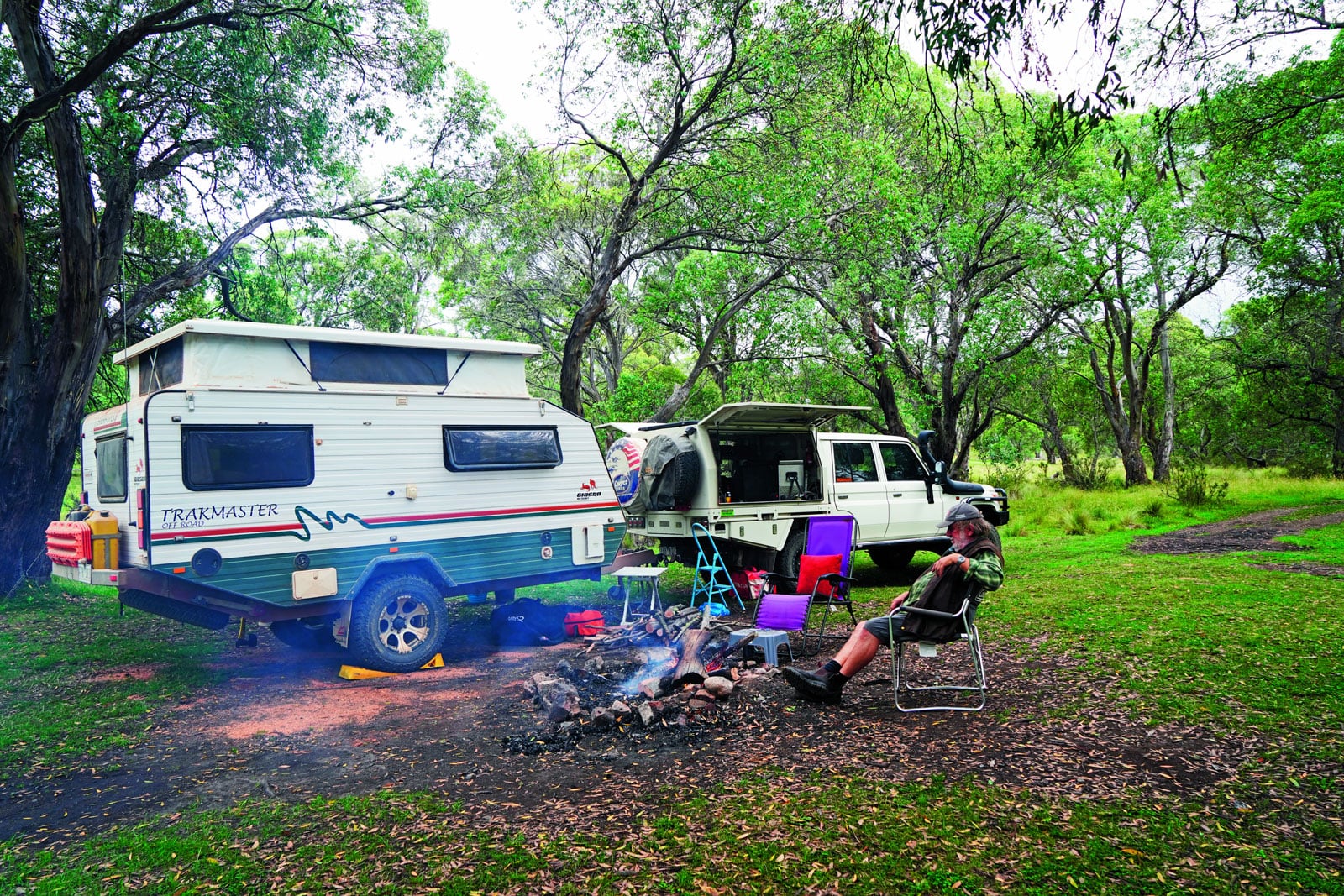 Two campers parked in a wooded campsite, with a campfire burning and a man relaxing in a lawn chair nearby.