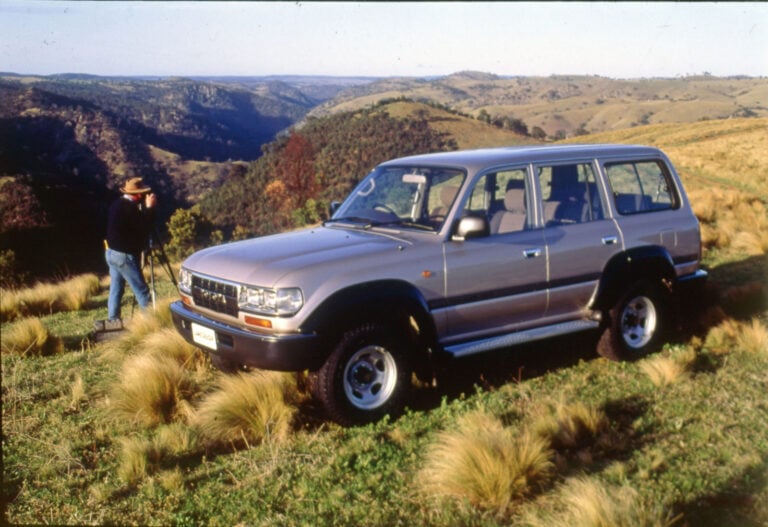 A man with a hat sets up a camera on a tripod beside a gray SUV on a grassy hillside overlooking a canyon landscape.