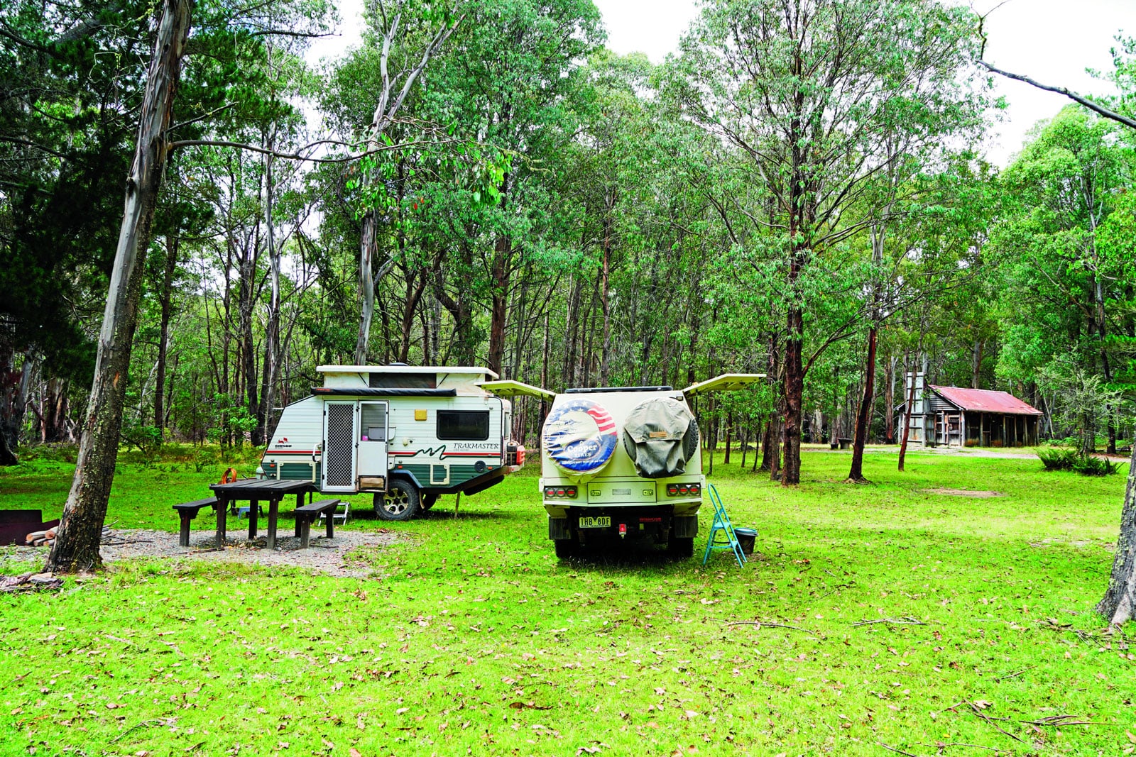 Campground with a white caravan and a green trailer parked among tall trees; a wooden picnic table sits nearby on a grassy clearing.