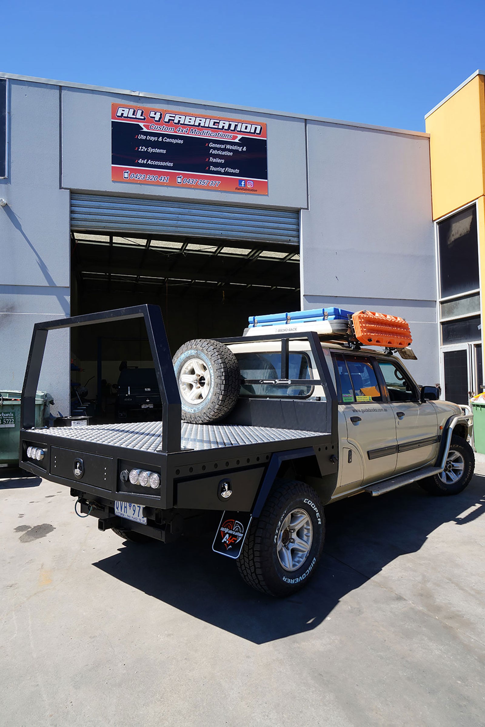 Modified off-road pickup with a heavy-duty black flatbed on a trailer outside an industrial shop; sign reads 'All 4 Fabrication' in the background