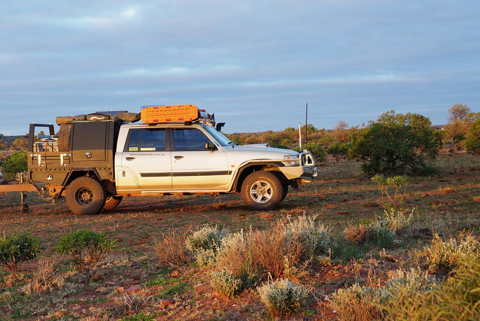 White 4x4 off-road vehicle equipped with roof rack, orange storage case, and camping gear in a desert landscape at sunset/near dusk.