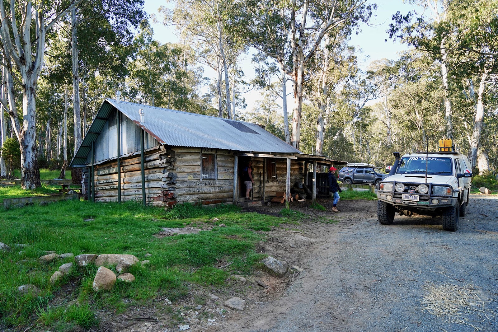 Rustic log cabin in a forest clearing with two off-road vehicles nearby and people entering the cabin.
