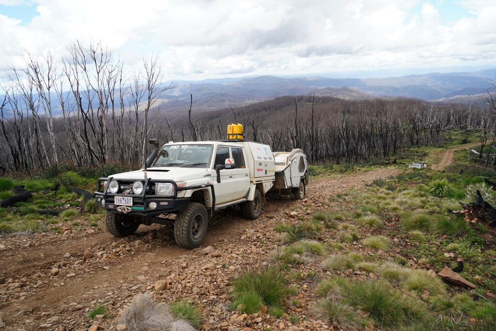 White 4x4 utility truck with a trailer on a rocky mountain track through a scorched forest. (Informative)