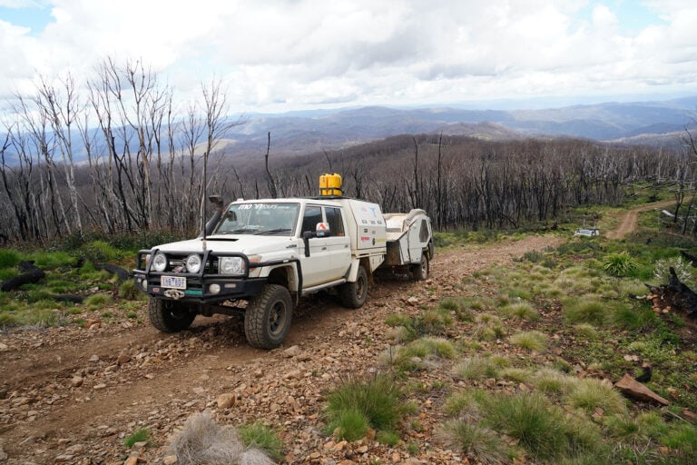 White 4x4 utility truck with a trailer on a rocky mountain track through a scorched forest. (Informative)