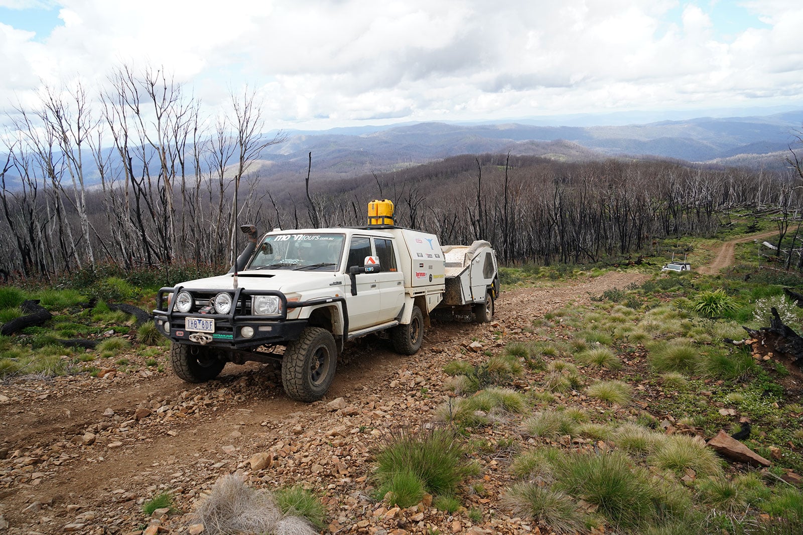 White 4x4 utility truck with a trailer on a rocky mountain track through a scorched forest. (Informative)