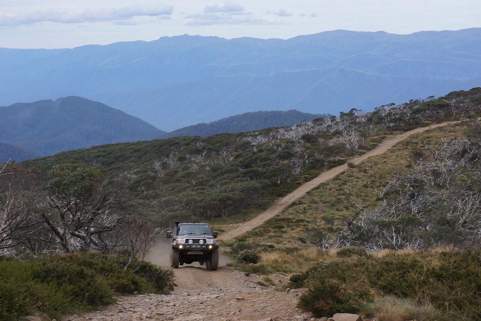 Off-road vehicle driving on a rocky dirt trail through scrub with distant blue mountains in the background.