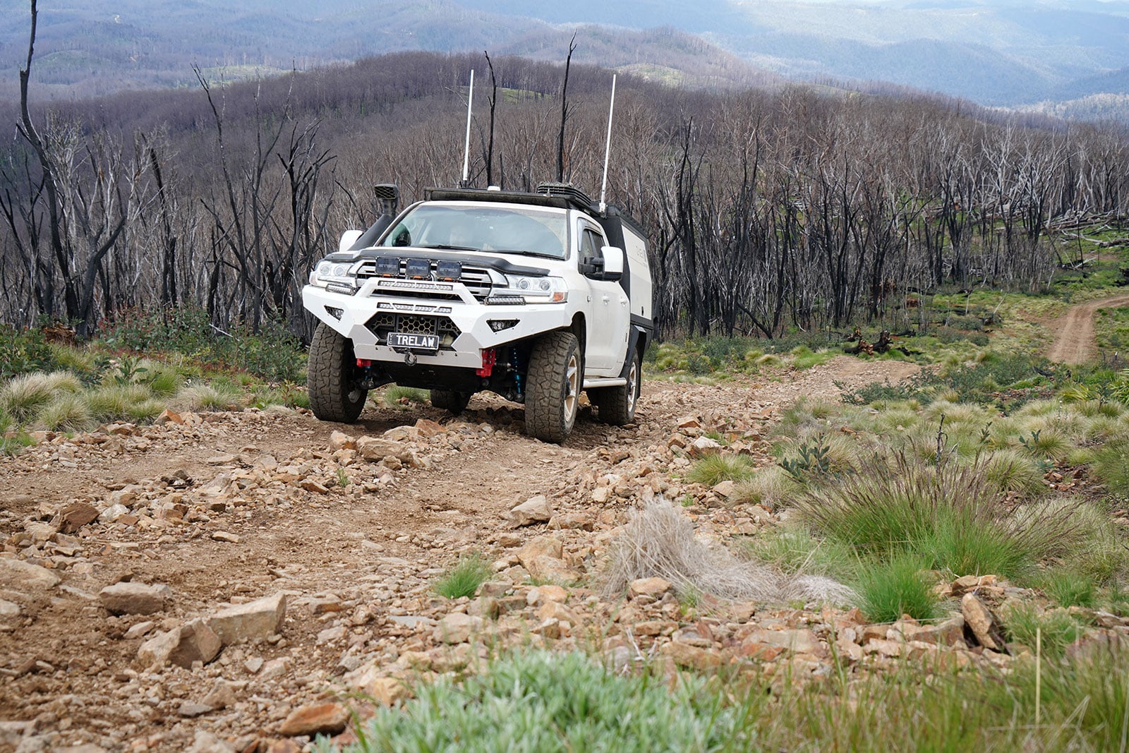 White off-road truck navigating a rocky dirt trail through a burned forest with distant mountains curving in the background.