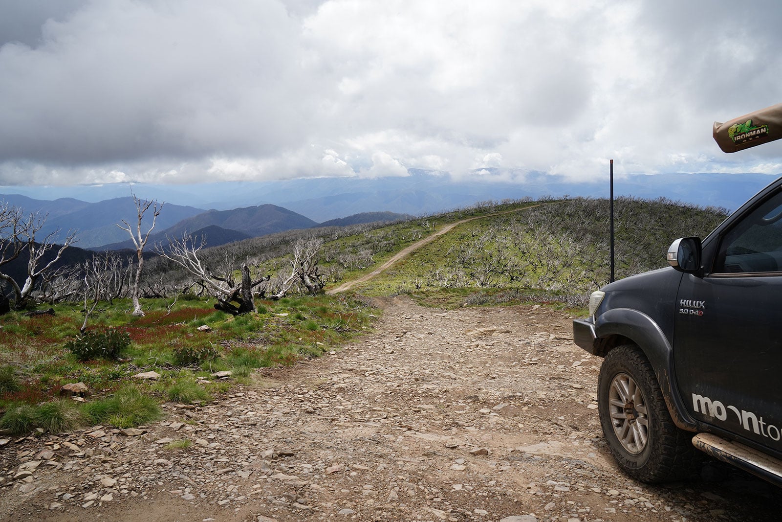 Rugged dirt road curling up a mountainside with leafless trees; a dark SUV parked on the right against a cloudy sky.