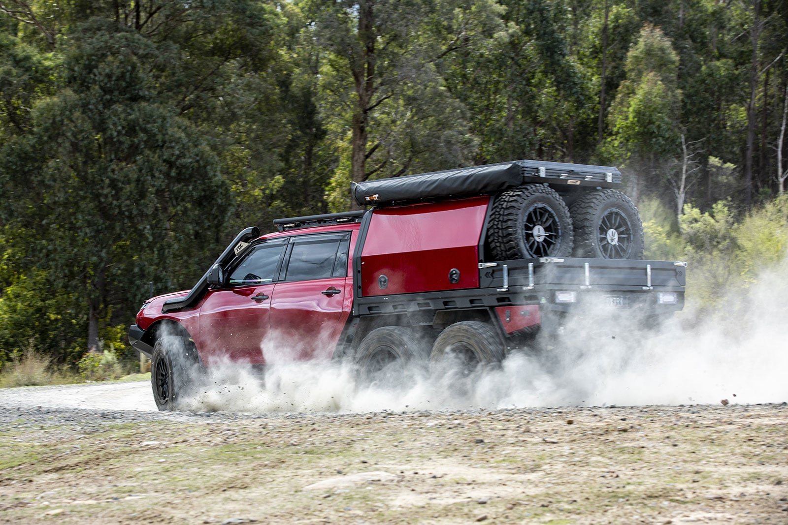 Red off-road pickup kicking up dust as it speeds along a dirt road, with two spare tires secured in the bed and a black canopy above the cab.