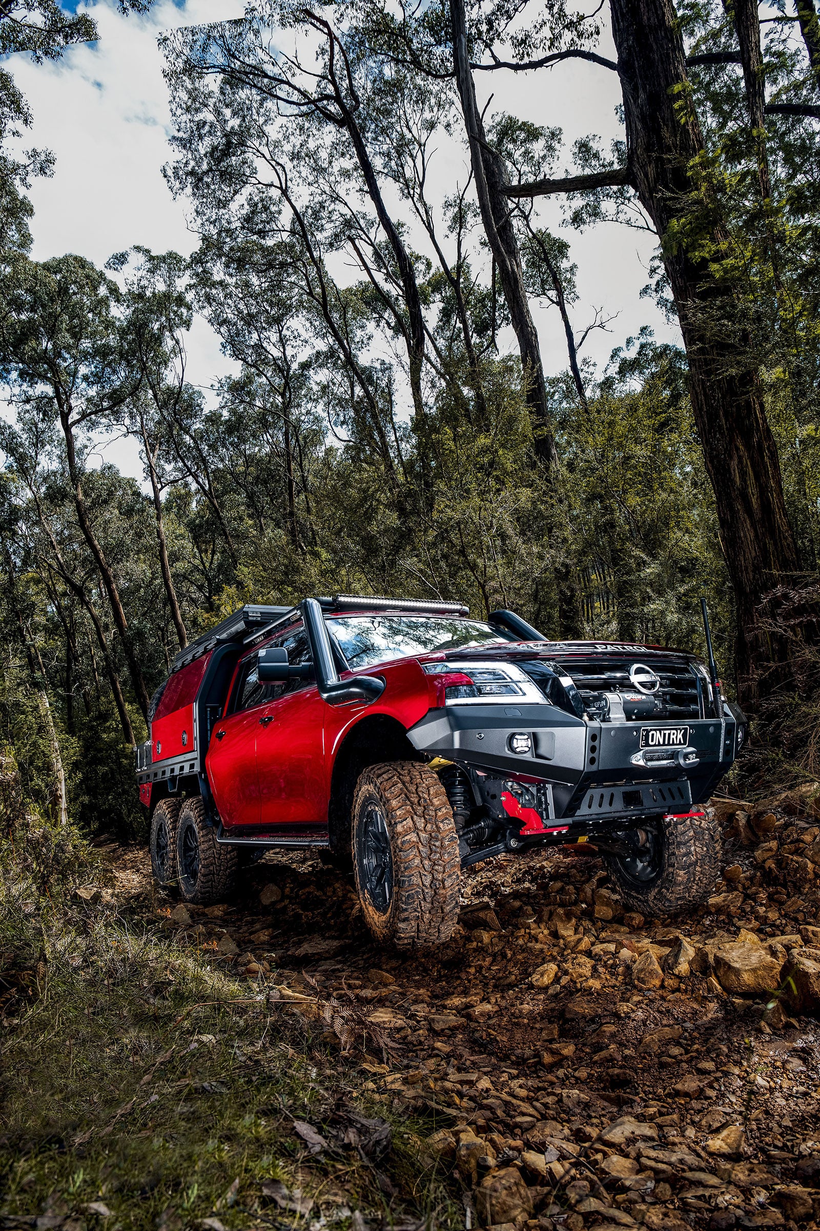 Red off-road pickup on a rocky forest trail with tall trees and blue sky peeking through.
