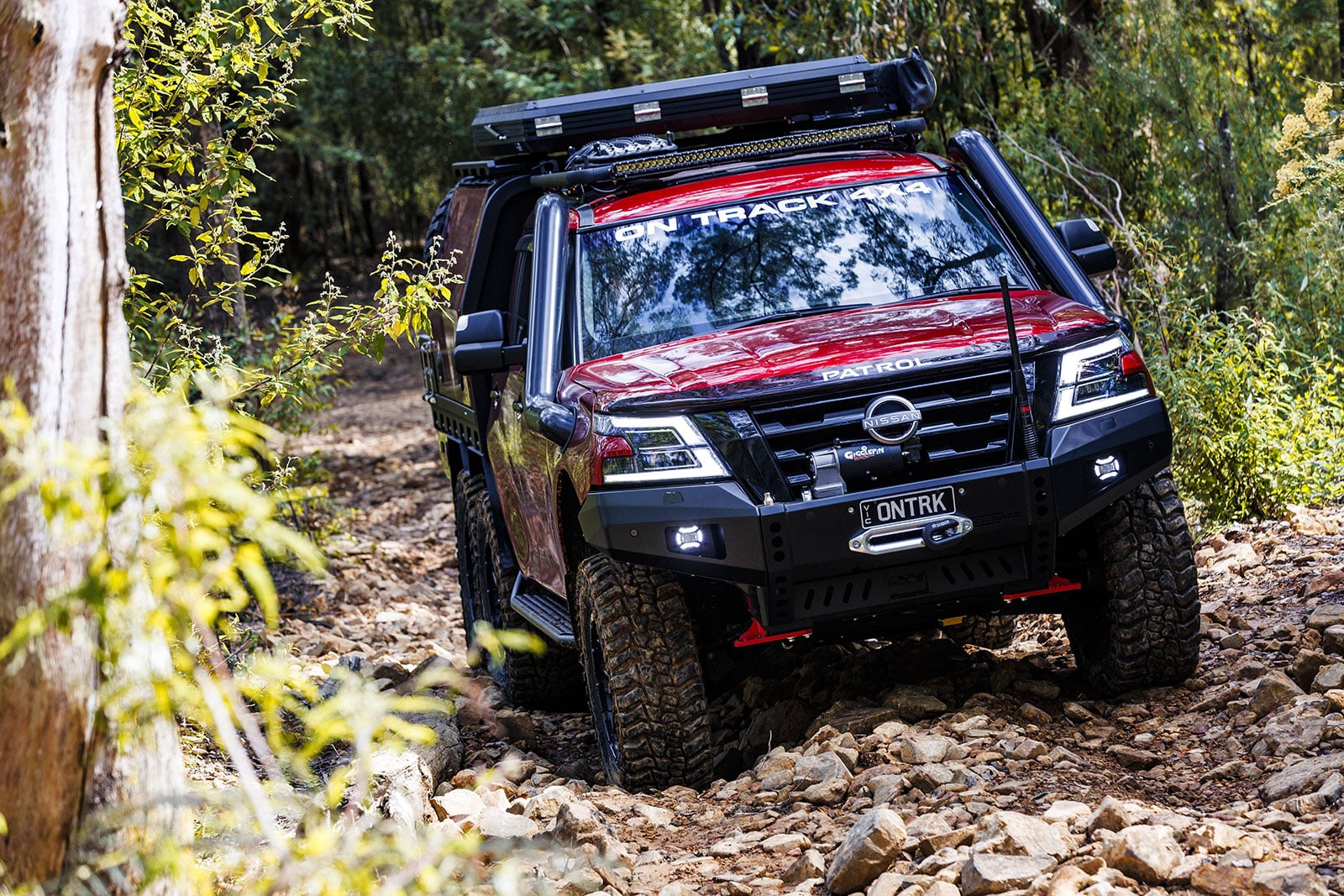 Red Nissan Patrol off-road SUV navigating a rocky forest trail with a roof rack and LED bar on top.