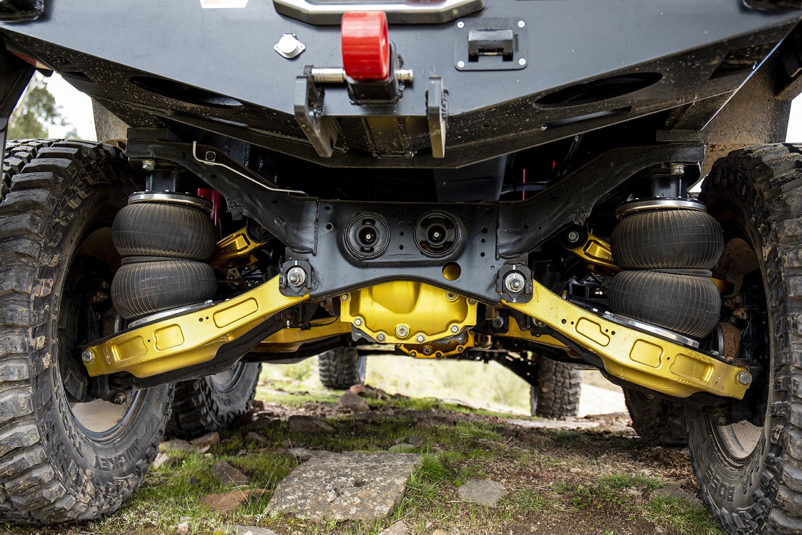 Underbelly of an off-road vehicle showing dual coilover air suspension, gold control arms, and rugged tires visible from below.