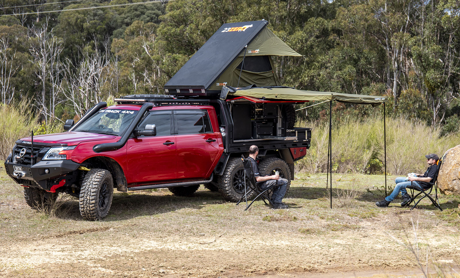 Red off-road pickup with a rooftop tent and extended awning camping setup in a grassy clearing, two people seated in chairs nearby.