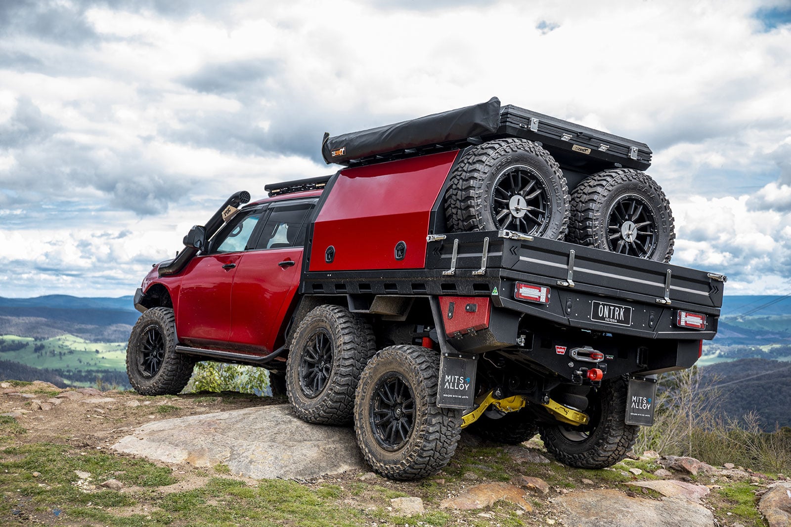 Red 4x4 pickup with a spare-tire rack on a rocky hill, with a scenic valley in the distance.