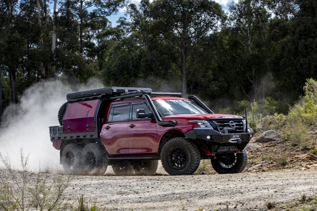 Red six-wheel off-road truck kicking up dust on a dirt trail in a forested area.