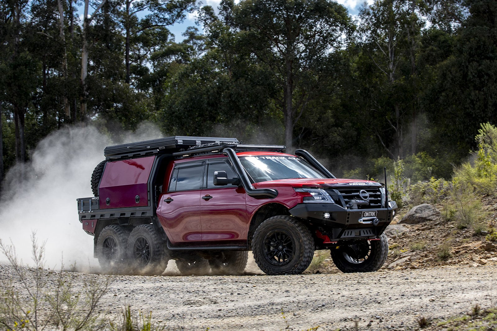 Red six-wheel off-road truck kicking up dust on a dirt trail in a forested area.
