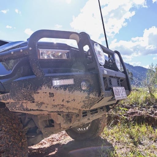 Front view of a mud-splattered off-road SUV navigating a rocky dirt trail with a blue sky and mountains in the background, bumper guard and lights visible.