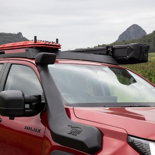 Red Toyota Hilux pickup with a black roof rack and cargo box, parked in a grassy field with hills and a distant mountain.