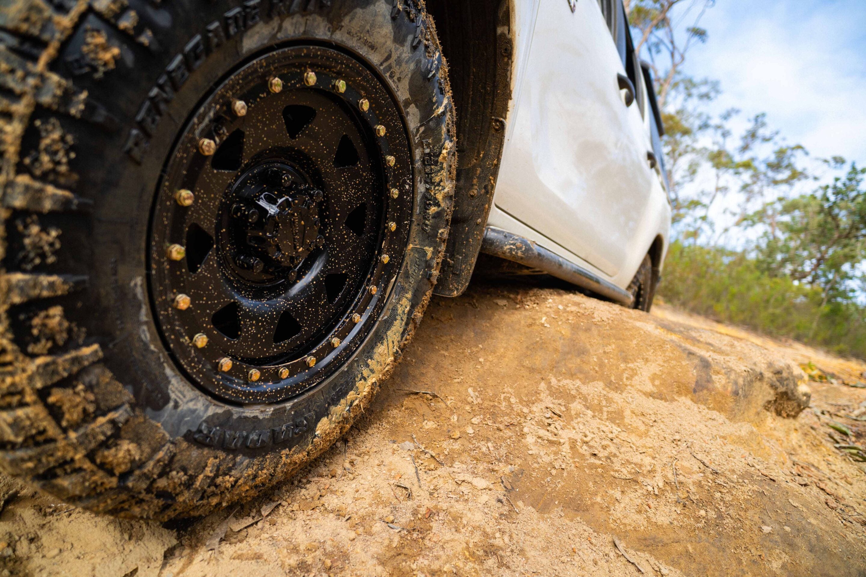 Close-up of a muddy off-road tire and wheel on a dusty dirt trail, white vehicle body in the background.