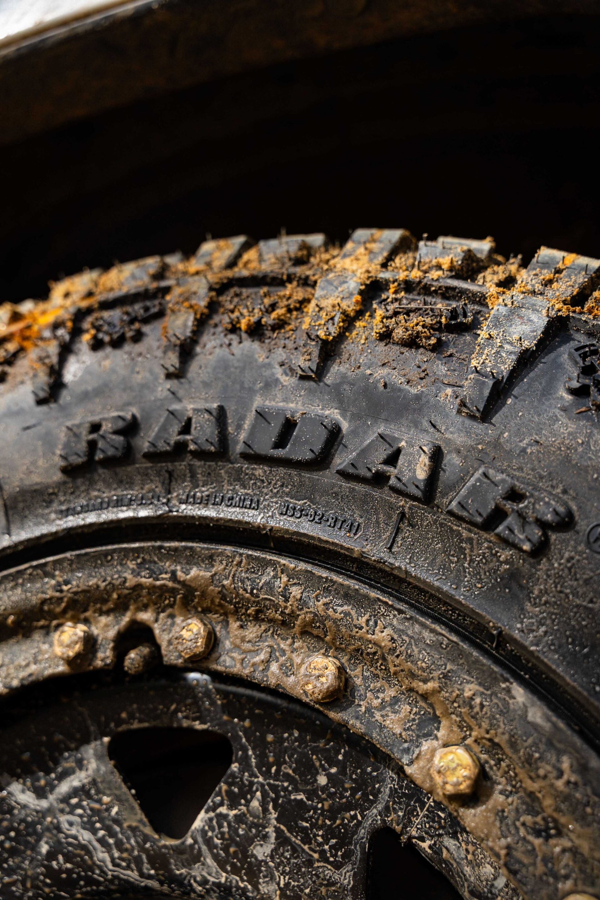 Close-up of a dirty off-road tire with mud caked in the tread blocks and small rocks stuck in the grooves.
