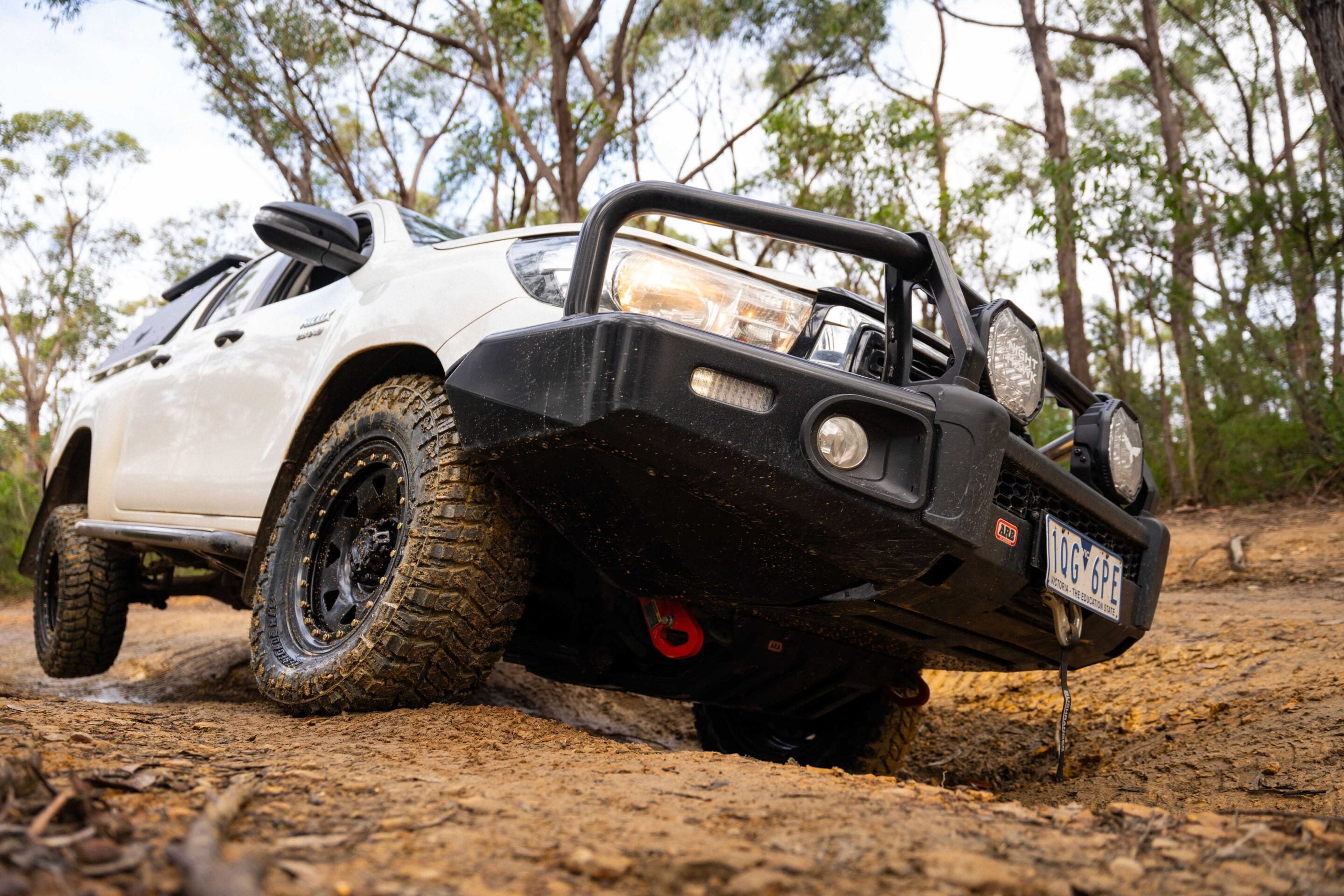 White off-road SUV with a heavy bull bar and muddy tires on a dirt trail among trees in a forest.