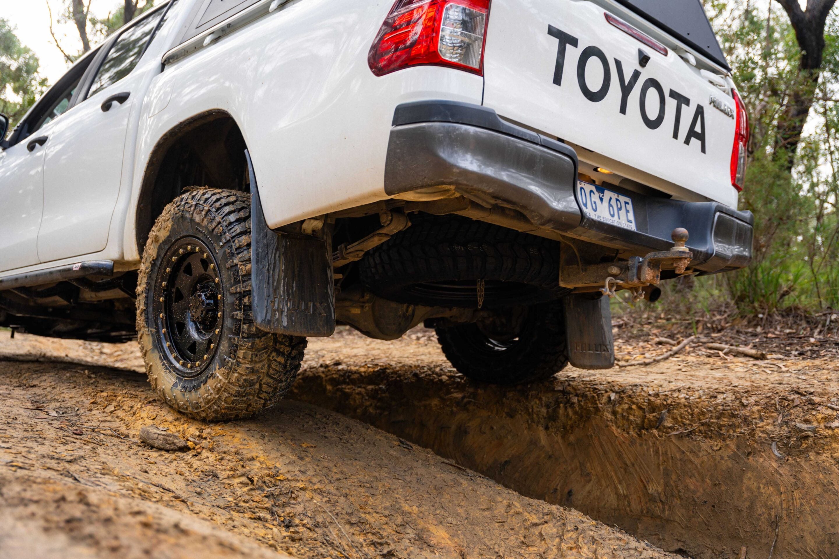 White Toyota pickup on a muddy off-road trail with the rear end elevated over a rut