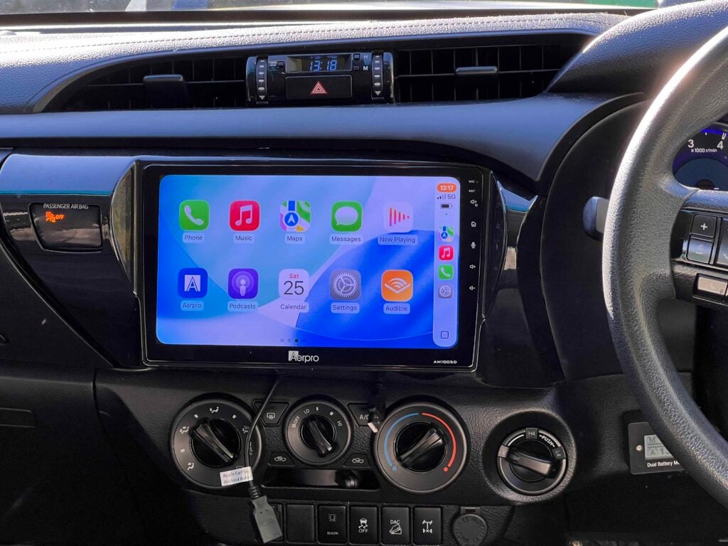 Dash panel with a large touchscreen showing iPhone apps (Phone, Music, Maps) and climate controls beneath, in a right‑hand drive car center console