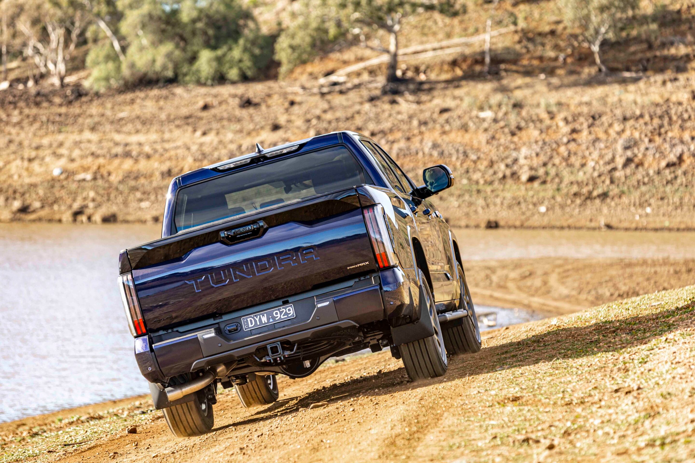 Blue Toyota Tundra pickup tilted on a dirt slope beside a body of water, hills in the background