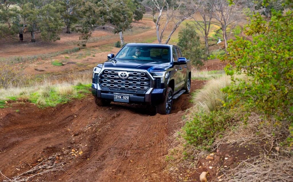 Blue Toyota pickup off-roading on a dirt trail through a grassy, hilly landscape.