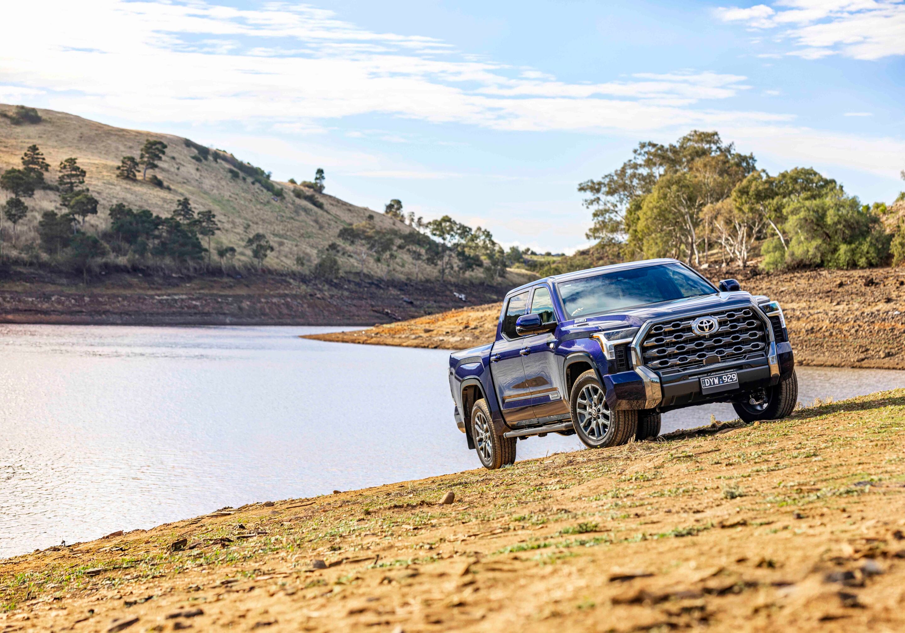 Blue pickup truck parked on a cracked, dry shoreline beside a calm lake with hills and trees in the background.