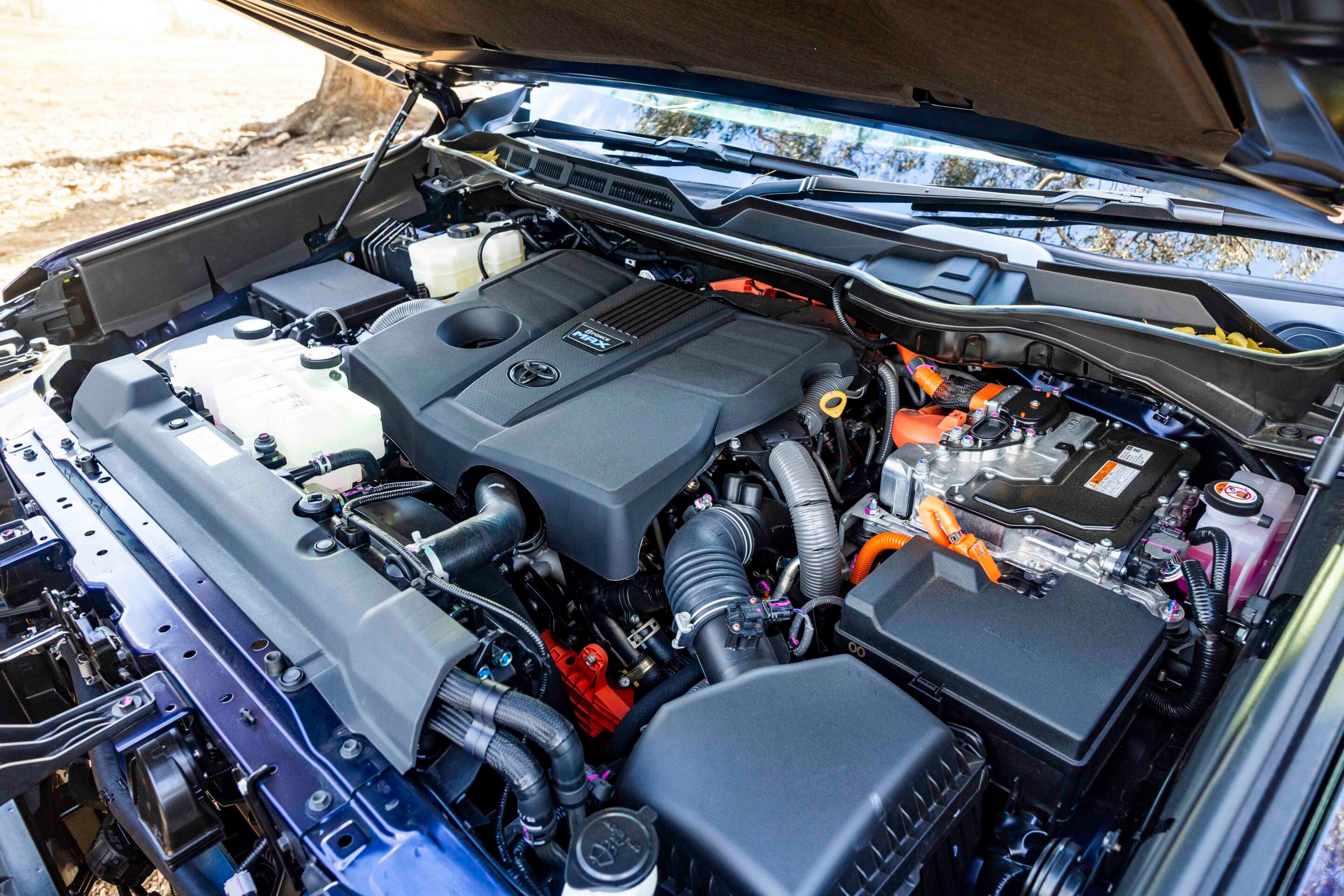 Open car hood showing the engine bay with battery, coolant reservoir, and various hoses and wires visible under the windshield area, outdoors.
