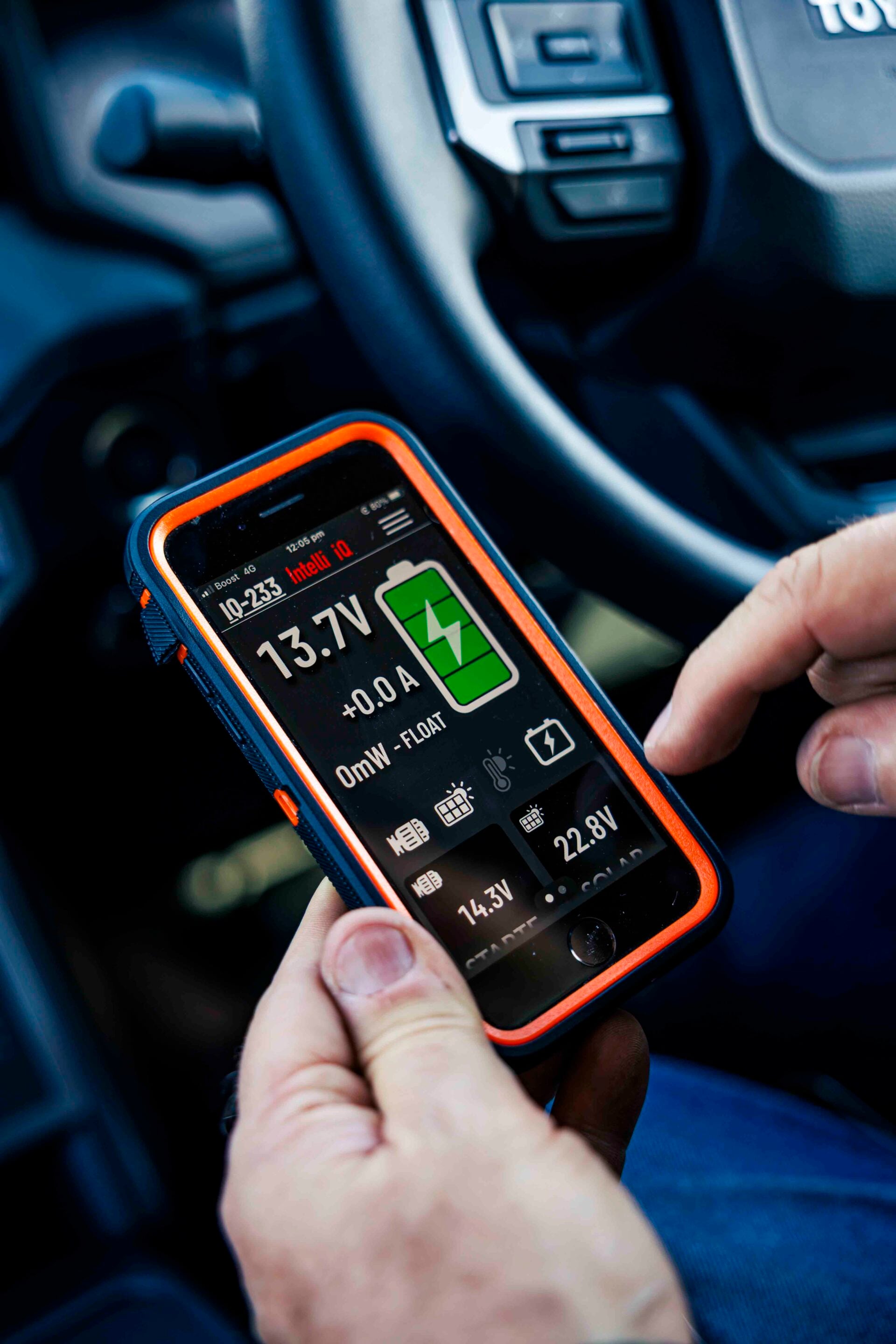 Close-up of a hand holding a rugged automotive battery tester in a car, showing 13.7V and a green battery icon.