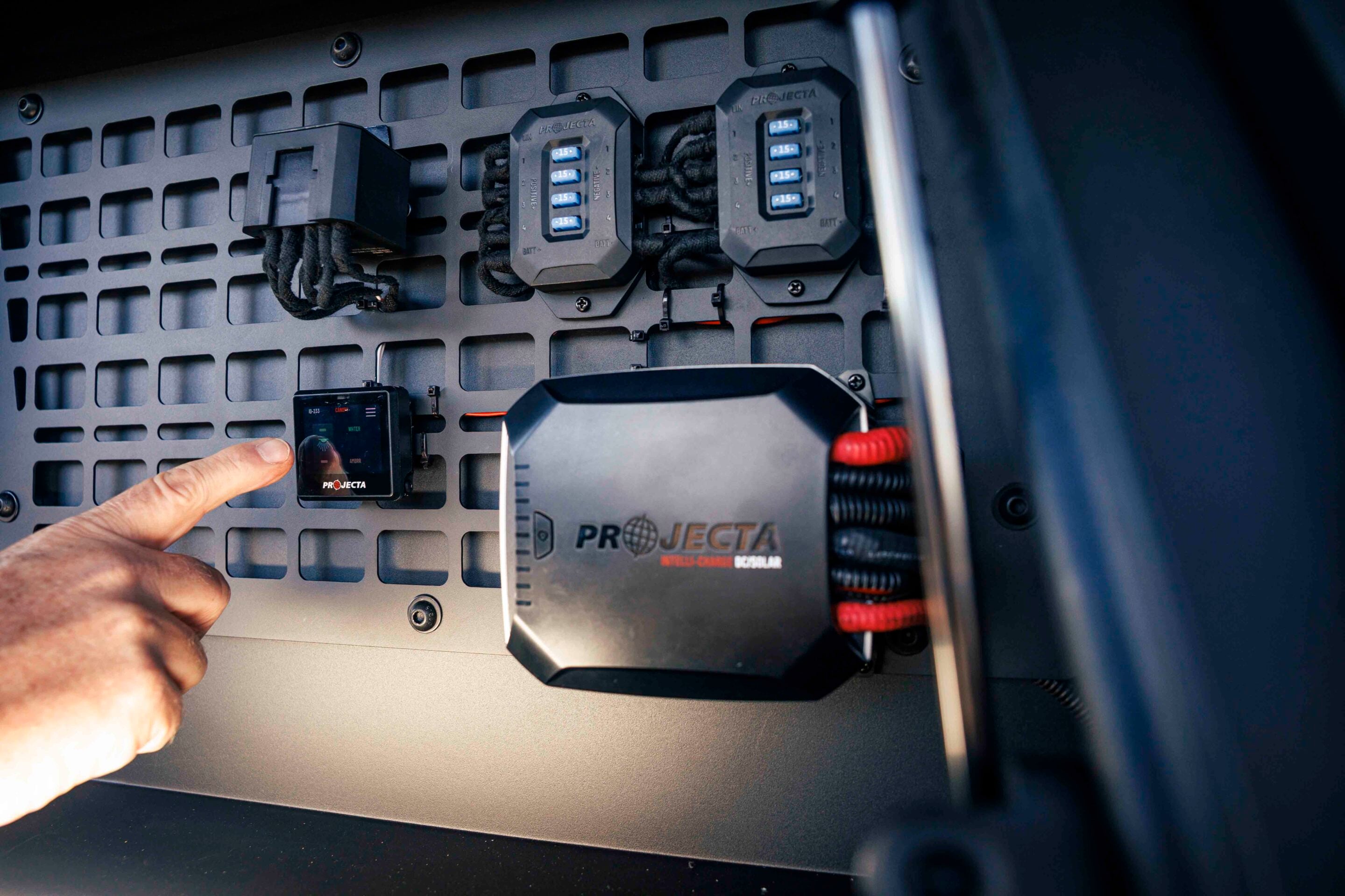 Close-up of a hand pointing at a PROJECTA installation with blue status indicators on a perforated panel inside a vehicle.