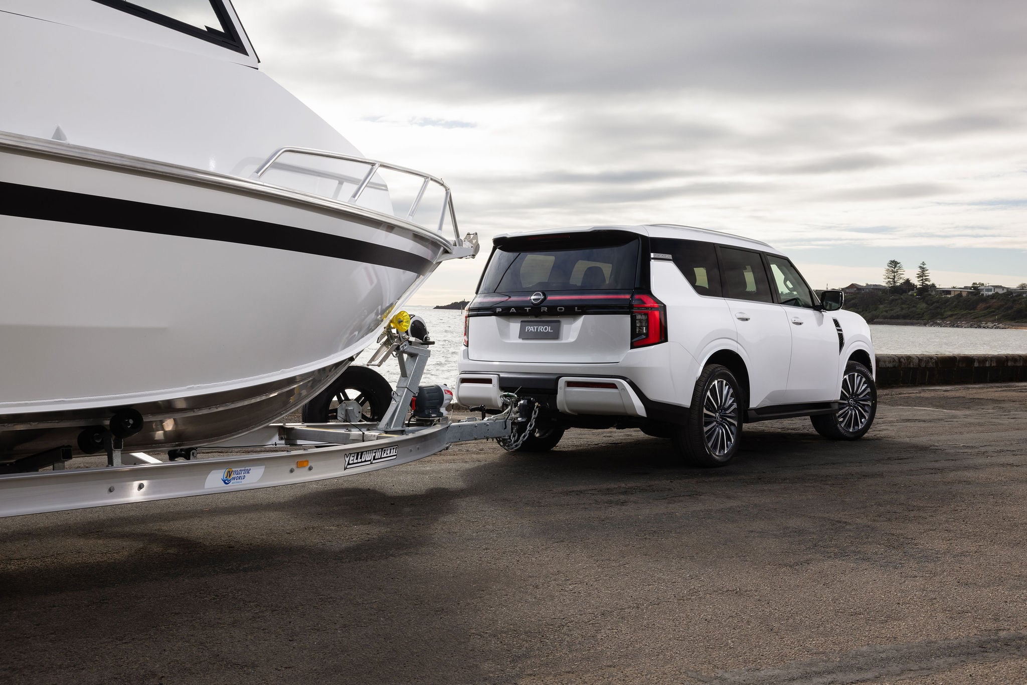 White yacht on a trailer hitched to a white Nissan Patrol SUV, parked at a marina with water in background and cloudy sky.