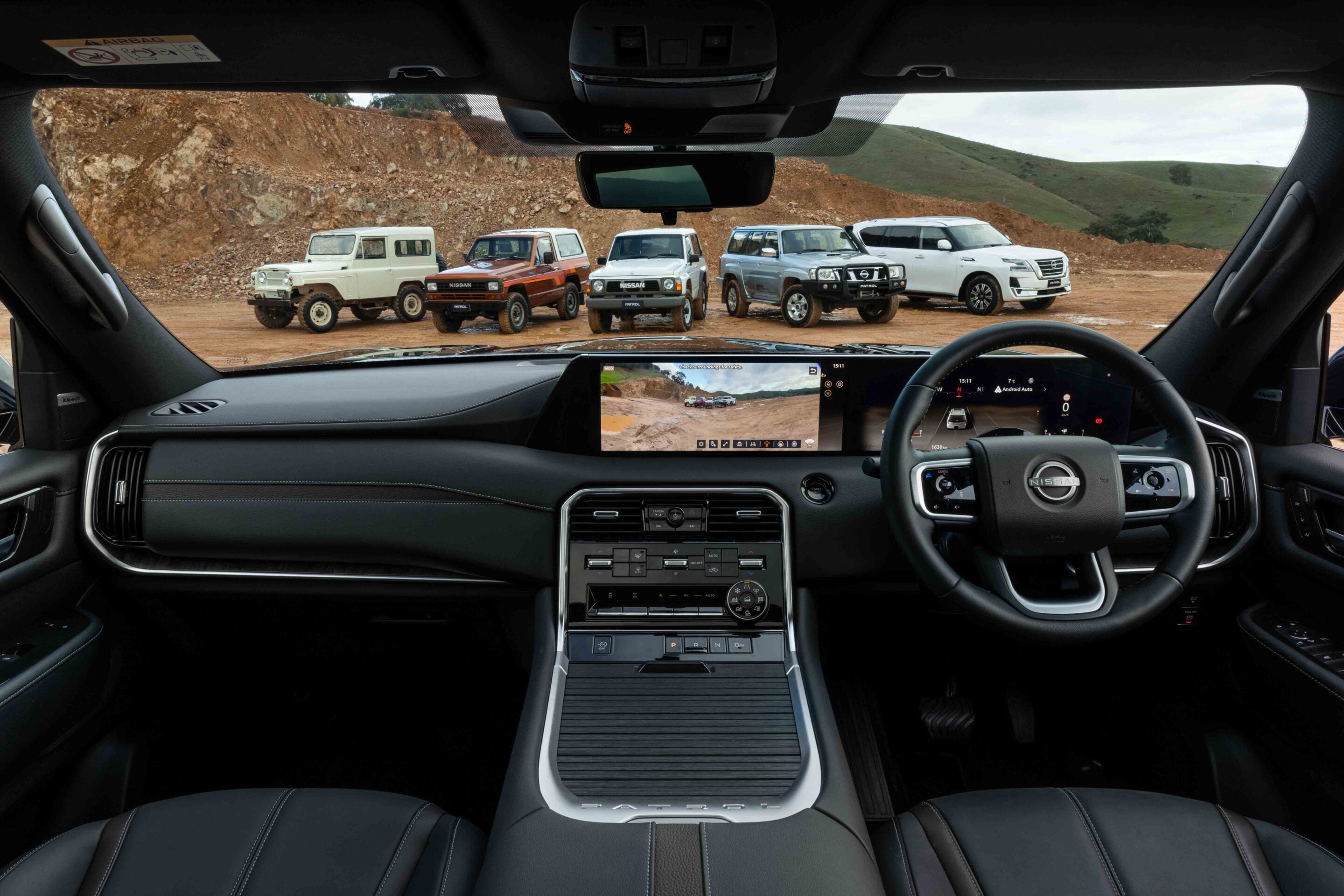 Interior view of a car cabin with a large central touchscreen and steering wheel, facing a row of off-road SUVs parked on dirt outside.