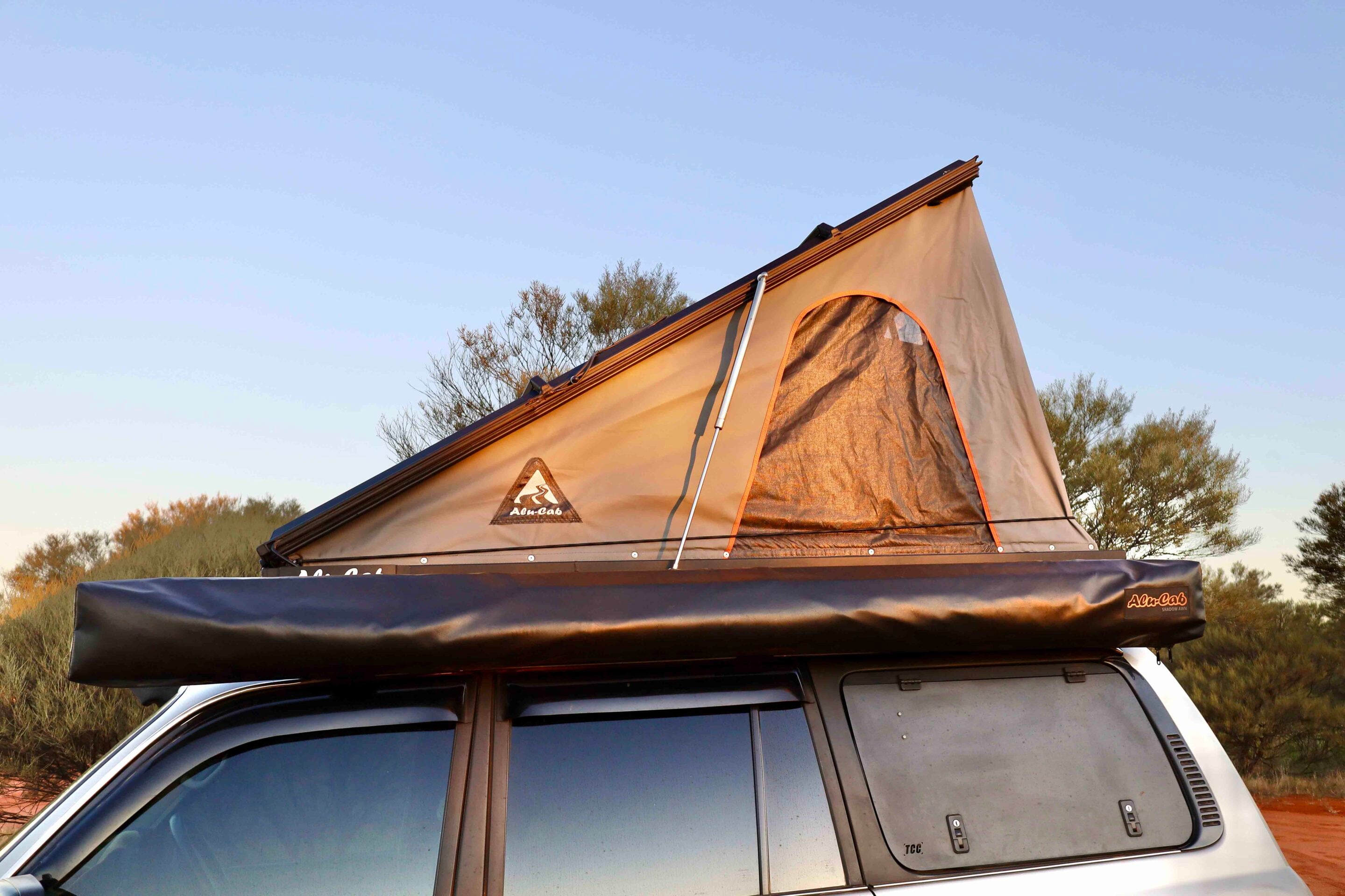 Tan rooftop tent mounted on a white SUV, set up outdoors with a clear blue sky and desert shrubs in the background, showing a side window and zipper opening edge at the tent peak.