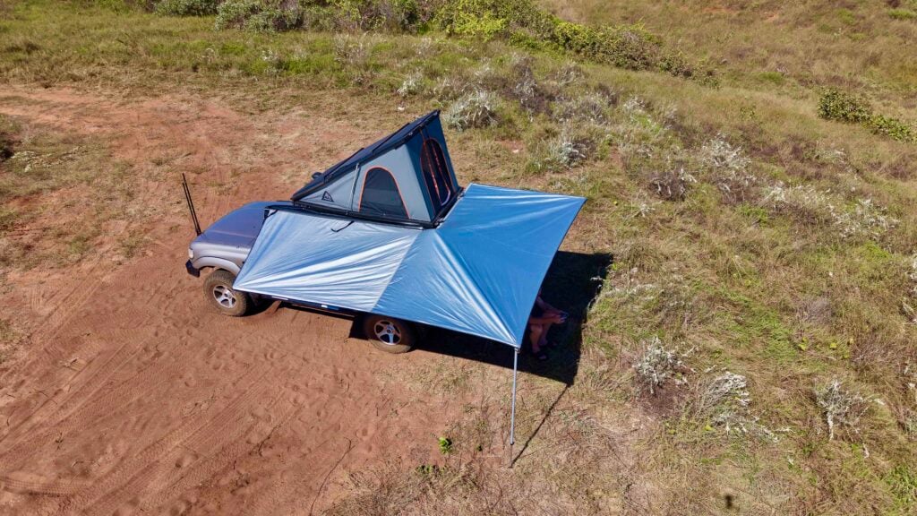 Overhead view of a silver SUV with a blue rooftop tent and blue canopy on a dirt trail in a grassy area court campsite setup.